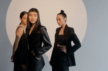 rês mulheres em pé, vestidas com roupas pretas elegantes, estão de frente para a câmera. Ao fundo, uma grande luz branca circular ilumina a cena.