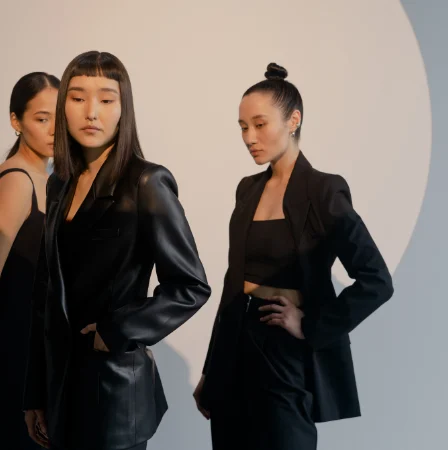 rês mulheres em pé, vestidas com roupas pretas elegantes, estão de frente para a câmera. Ao fundo, uma grande luz branca circular ilumina a cena.