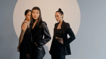 rês mulheres em pé, vestidas com roupas pretas elegantes, estão de frente para a câmera. Ao fundo, uma grande luz branca circular ilumina a cena.